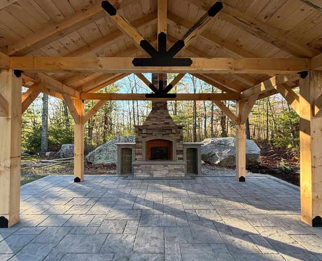 Wooden pavilion with a stone fireplace, on a gray stone patio. Forest in the background.