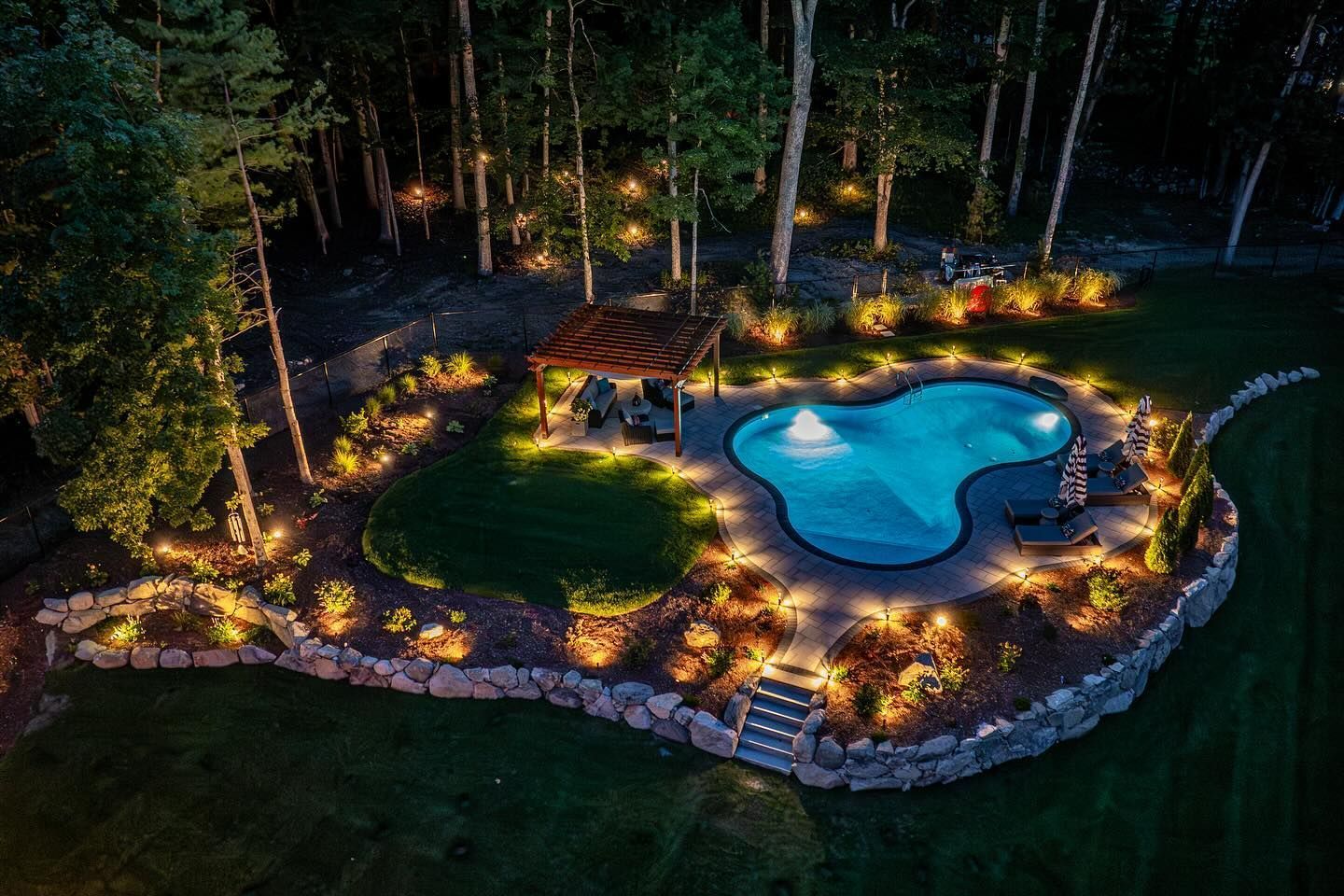 Nighttime view of a backyard with a pool, landscaping, and string lights illuminating a wooded area.