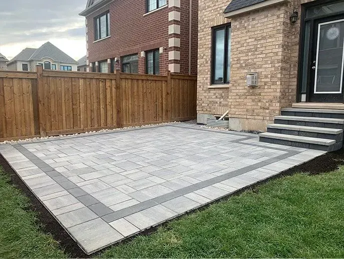 Patio with light gray pavers and dark gray border next to a house with brick siding, wooden fence, and green grass.