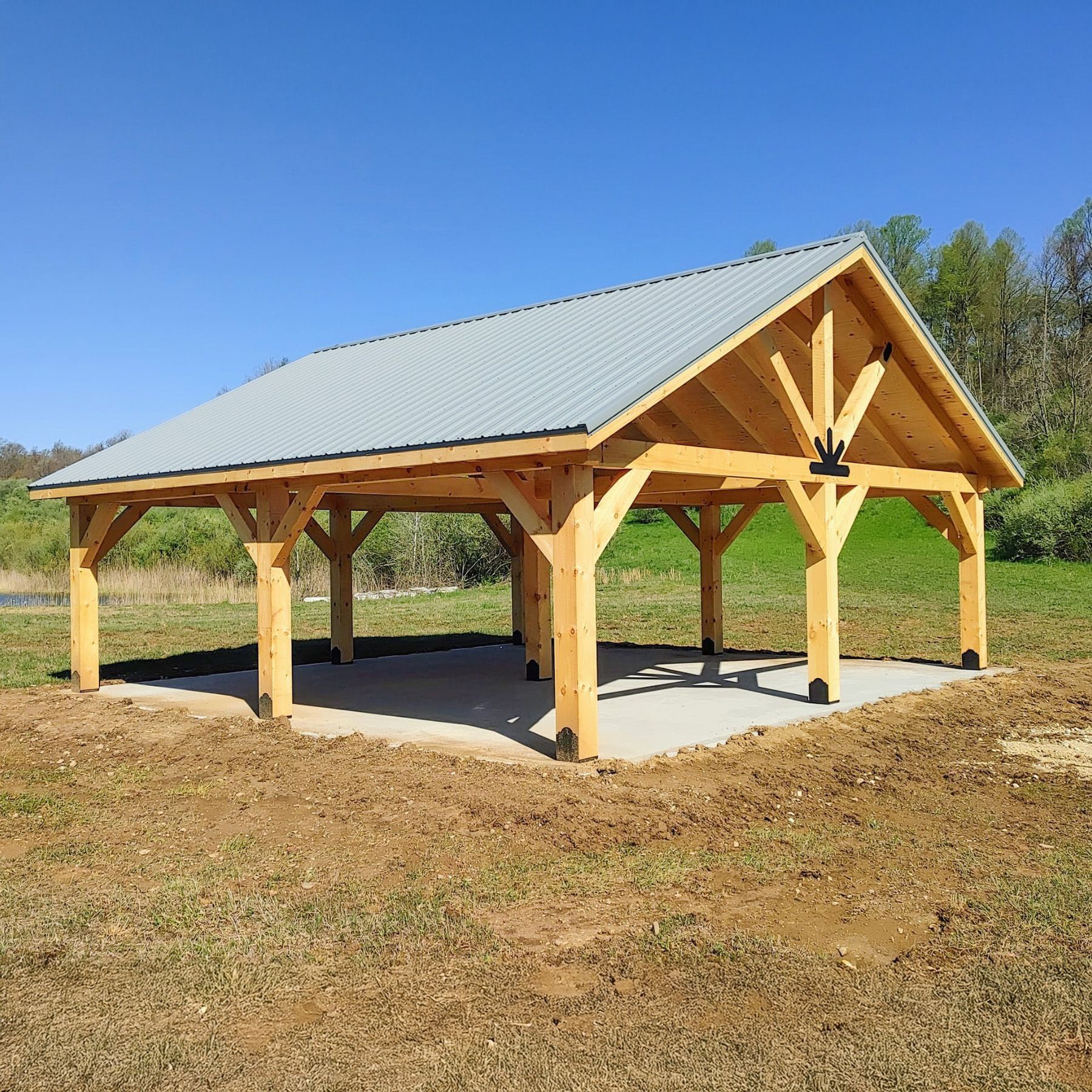 Wooden pavilion with gray metal roof on a concrete slab, set in a grassy field under a blue sky.