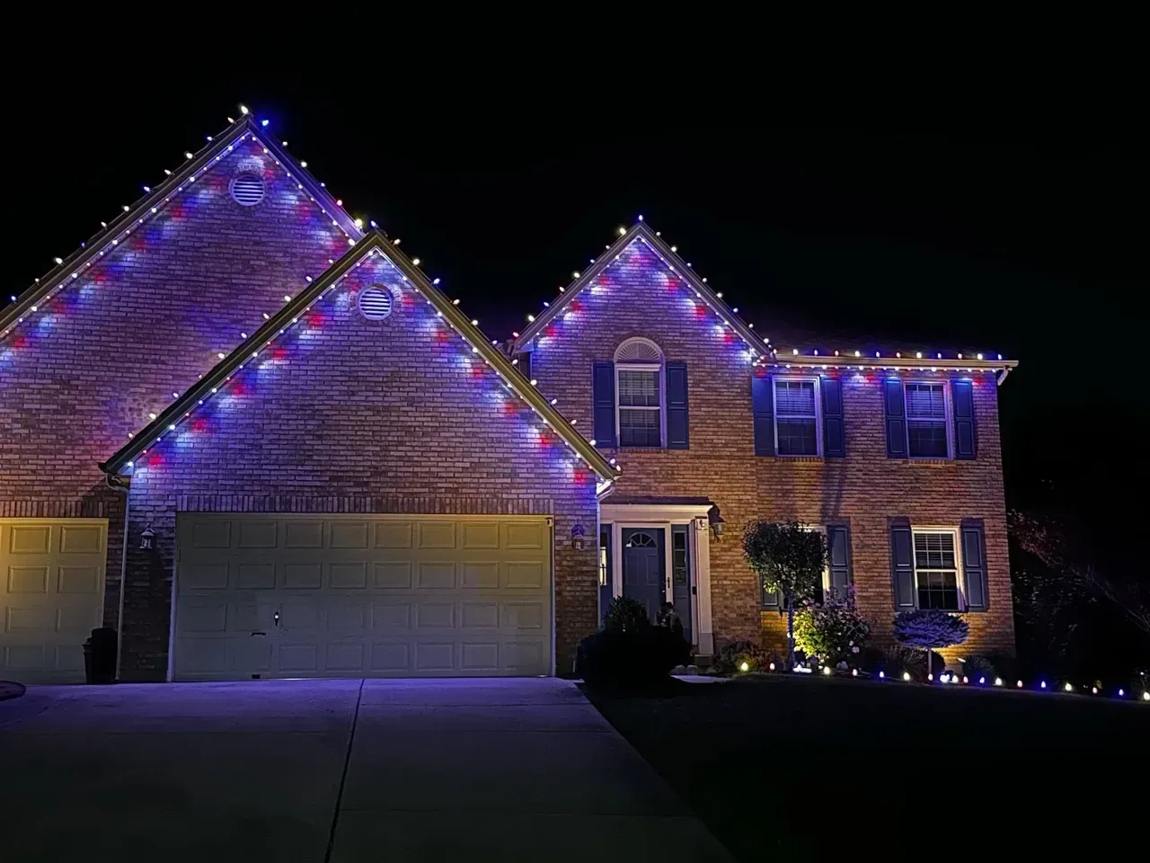 Two-story brick house illuminated with blue, red, and white Christmas lights along the roofline.