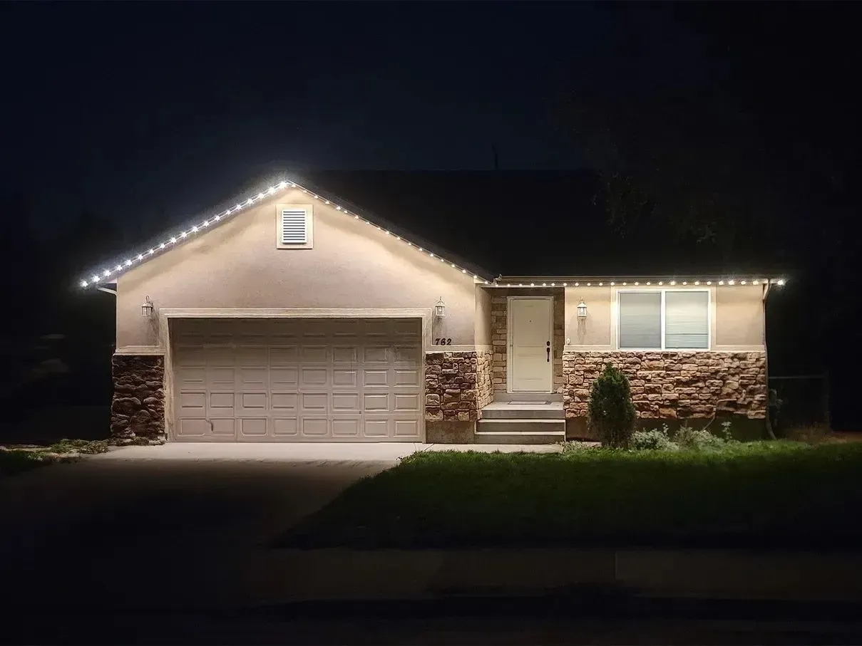 House at night, lit with white lights along the roofline. Beige siding, stone accents, garage.