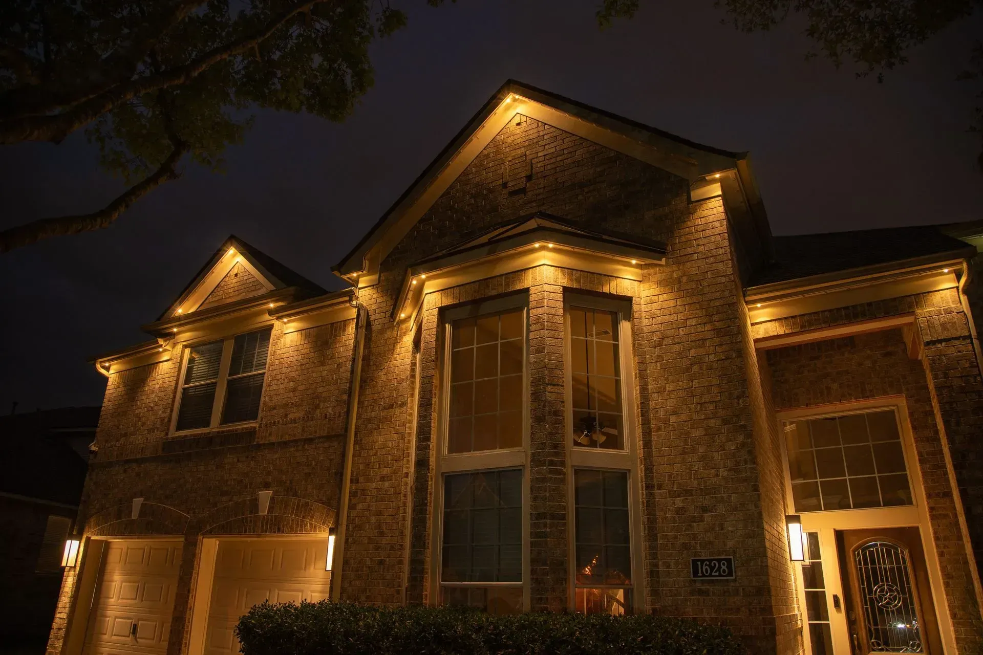 A two-story brick house illuminated at night with warm spotlights along the roof and facade.