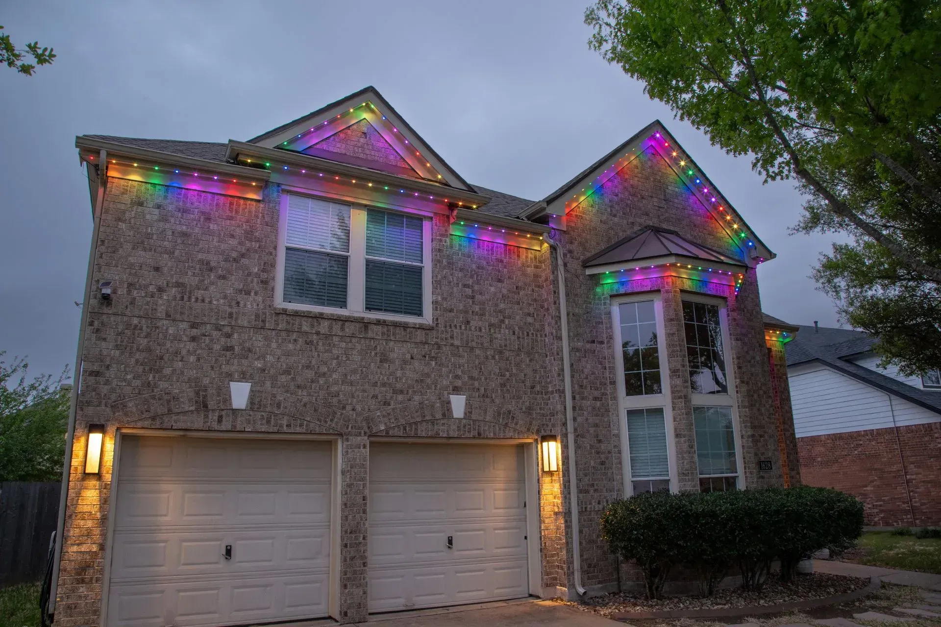 Two-story brick house decorated with colorful string lights along the roofline and above garage doors.