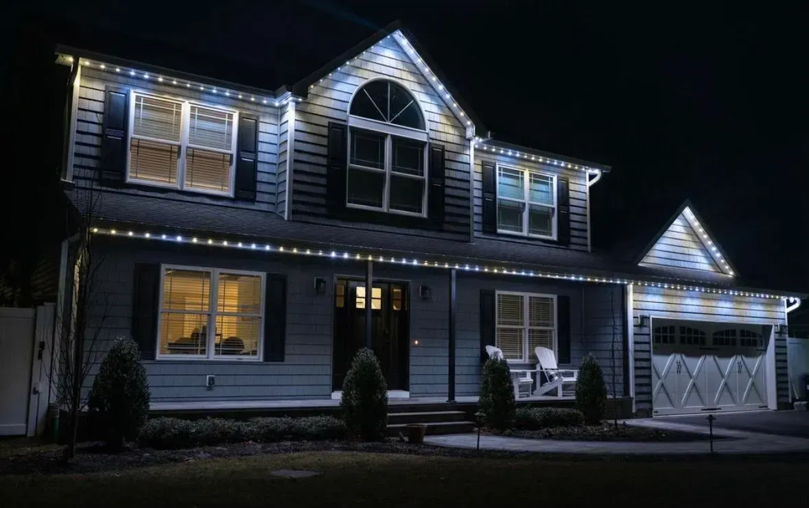 A two-story house at night, lit with white string lights along the roof and windows.