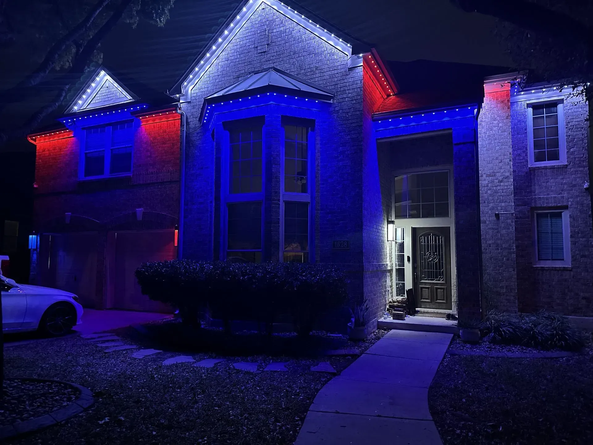 House with red and blue lights outlining the roof, windows, and pathway. A car is parked on the left.