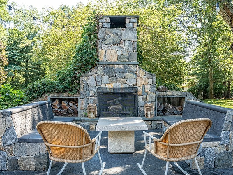 Outdoor stone fireplace with seating area, firewood storage, two wicker chairs, and a small table.