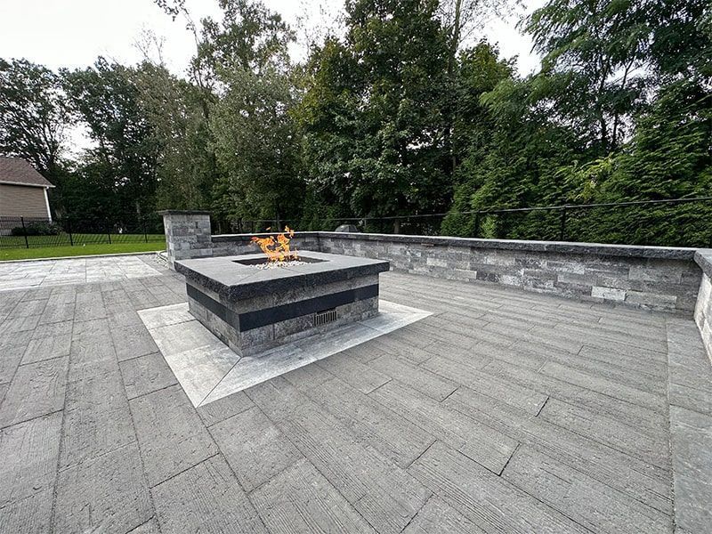Outdoor patio with lit fire pit; gray stone pavers and retaining wall; trees in the background.