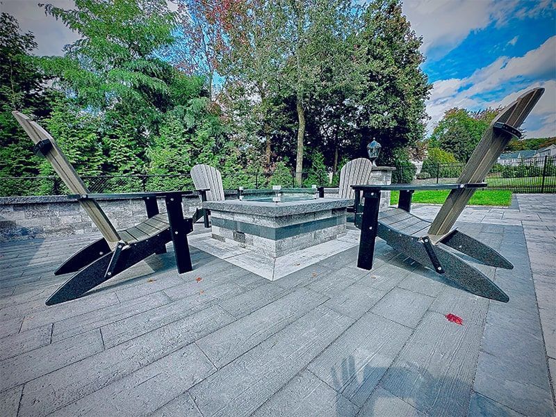 Two Adirondack chairs face a stone fire pit on a stone patio, trees in background.