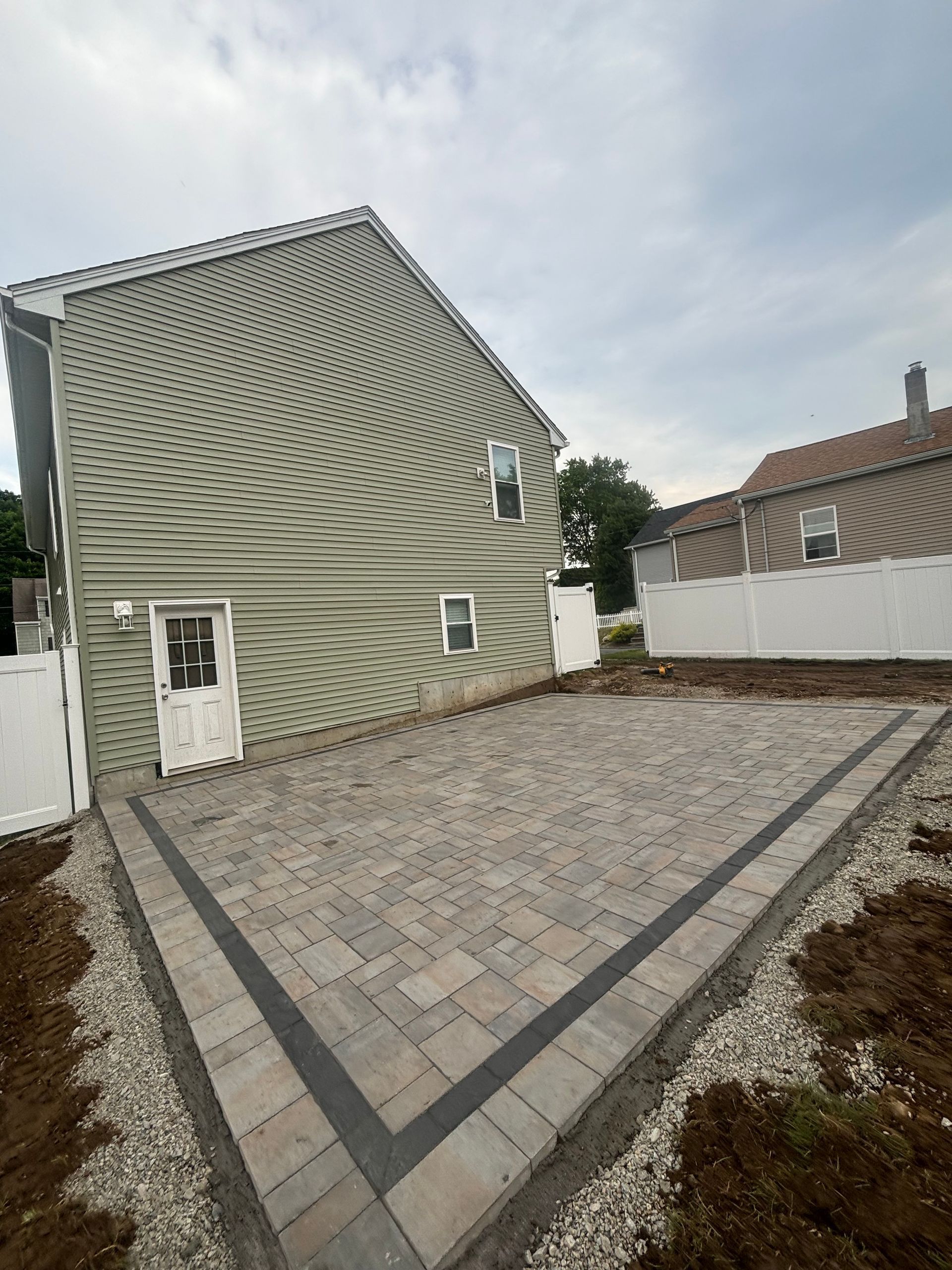 Backyard patio with gray pavers and a dark border, next to a green-sided house.