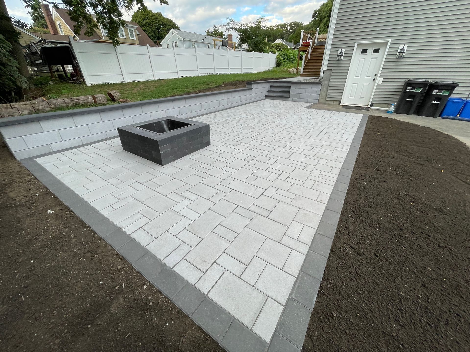 Paver patio with fire pit, bordered by dark pavers, retaining wall, and lawn.