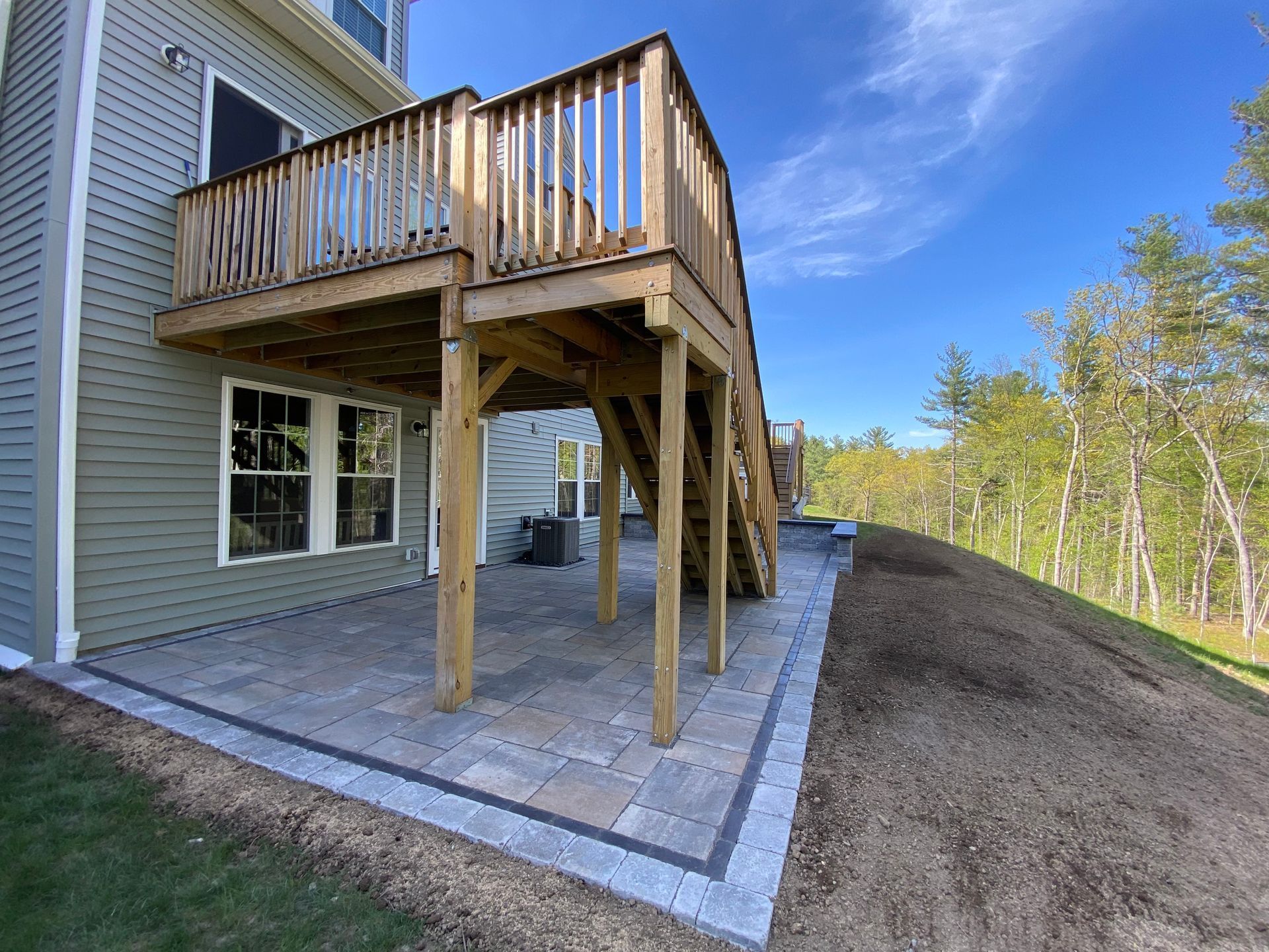 Deck with stairs over a brick patio, next to a green house, with a grass hill and blue sky.