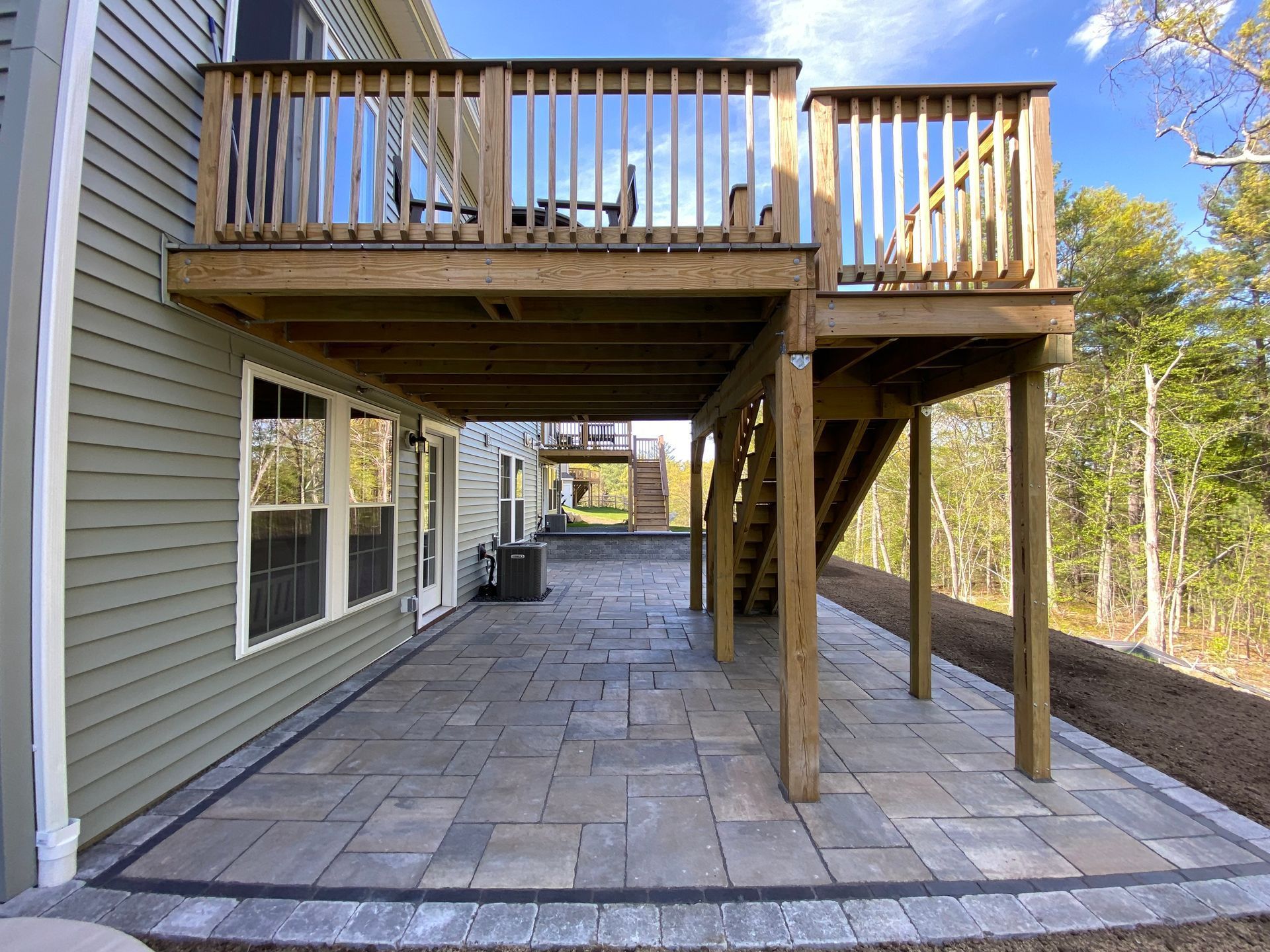Patio with a raised wooden deck above.  Stone pavers and green siding on a residential home under a blue sky.