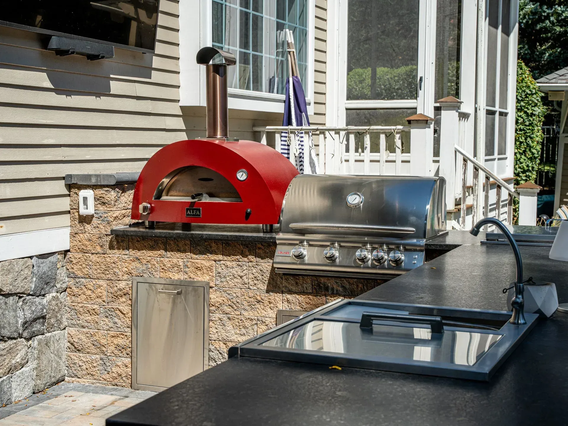 Red outdoor pizza oven and stainless steel grill on a stone countertop, near a sink and house exterior.