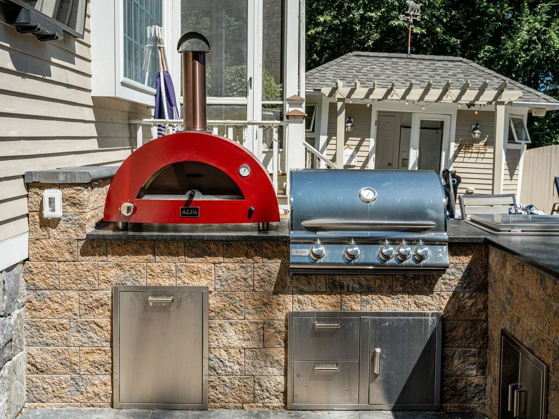 Outdoor kitchen with a red pizza oven, grill, and storage on a brick structure.