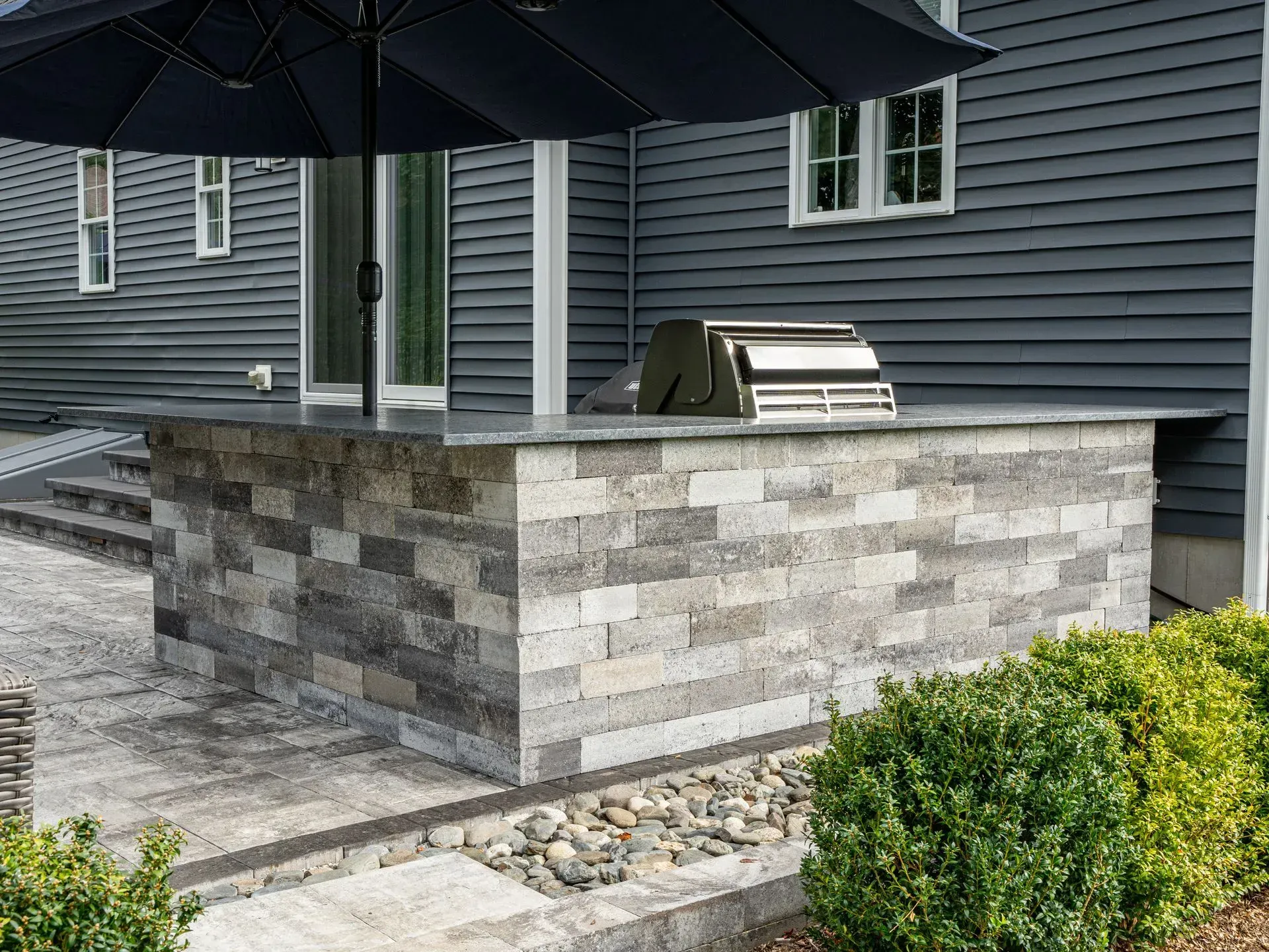 Outdoor kitchen with a gray brick facade, grill, and umbrella, set against a gray house.