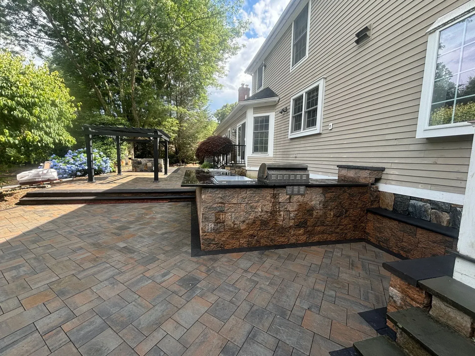 Brick patio with built-in grill and stone accents, next to a beige house, with steps leading to a pergola.