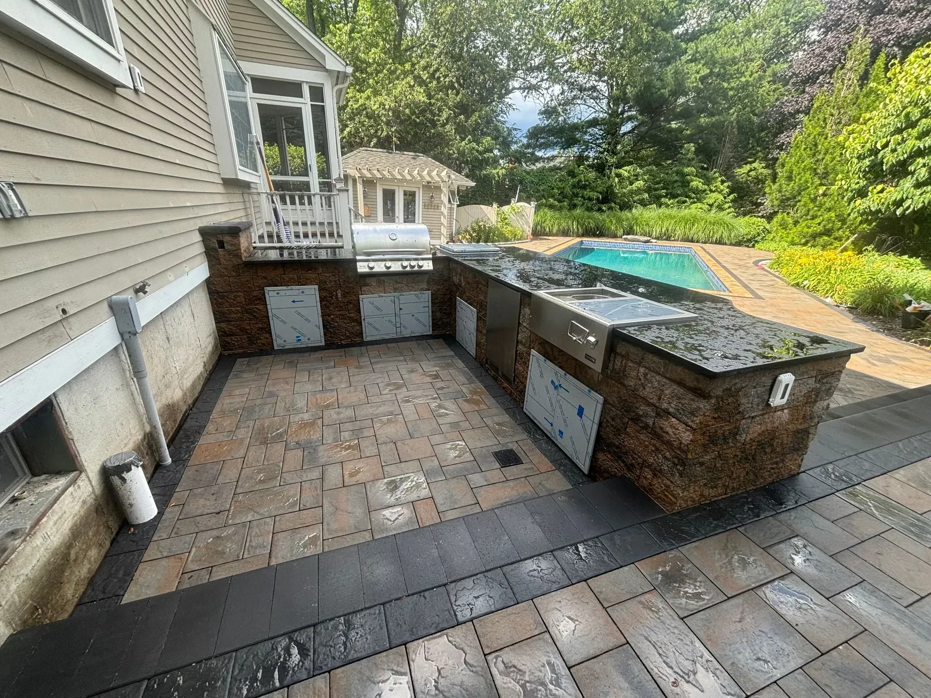 Outdoor kitchen with grill, pool, and stone patio. Beige siding, wooden structure, trees in background.