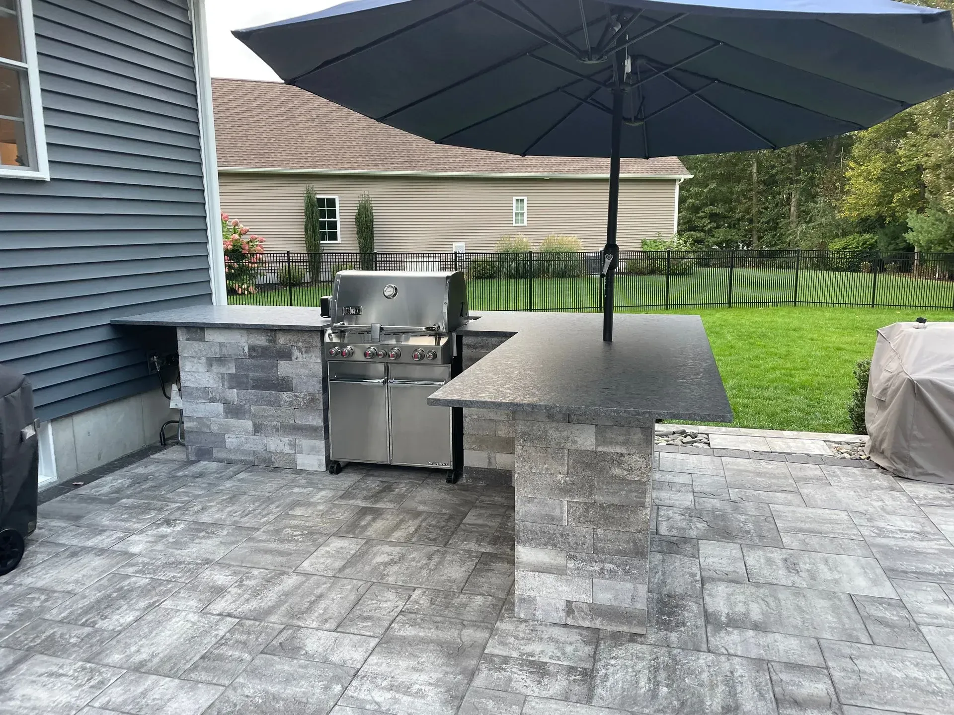Outdoor kitchen with a grill, countertop, and umbrella, set on a stone patio near a house.