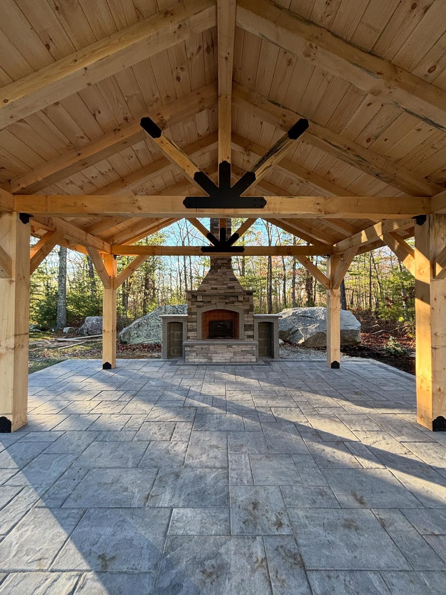 Wooden gazebo with stone fireplace and patio, surrounded by trees.