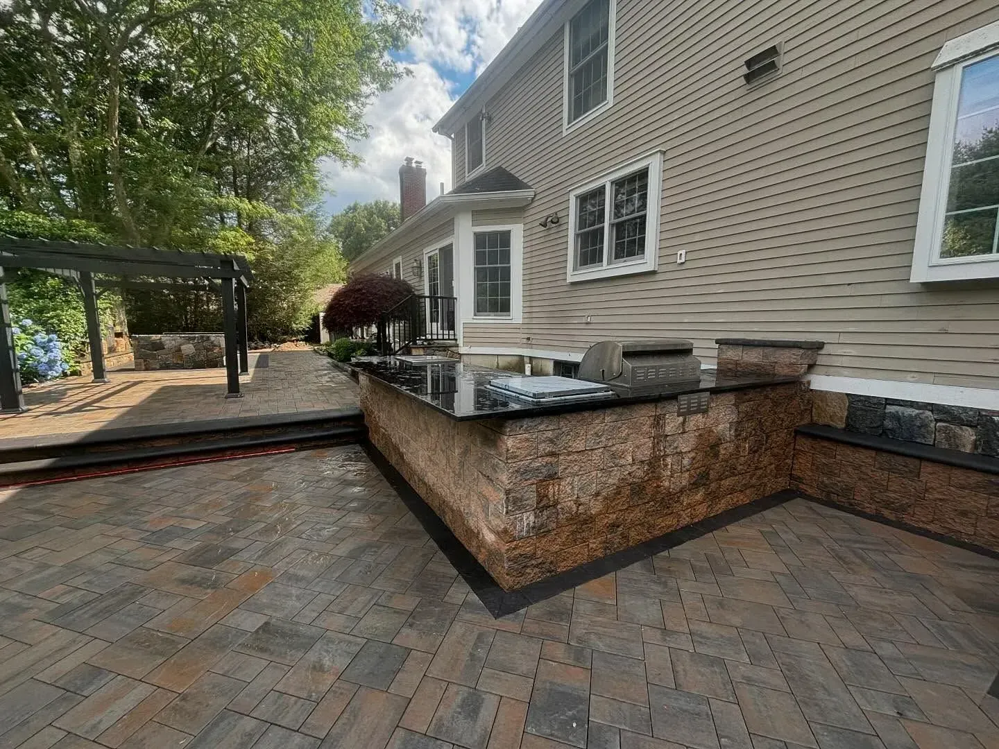 Outdoor patio with built-in grill and countertop against a two-story beige house. Brickwork, pavers, and dark pergola.