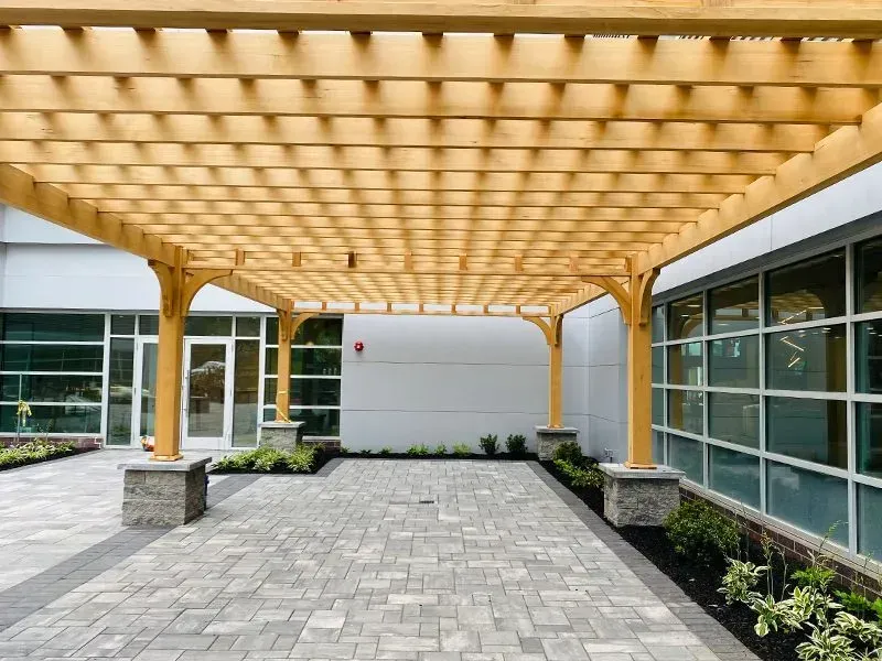 Wooden pergola over a gray brick patio, beside a building with large windows and a white wall.