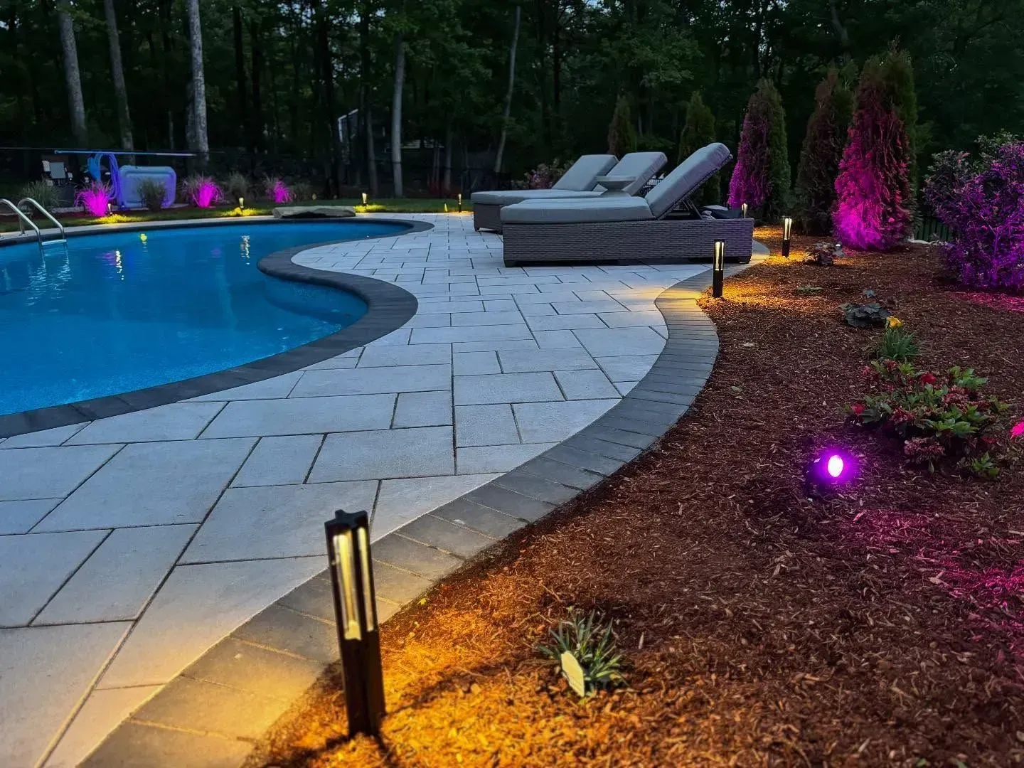 Poolside patio with stone pavers, lounge chairs, and landscape lighting in shades of orange and purple.
