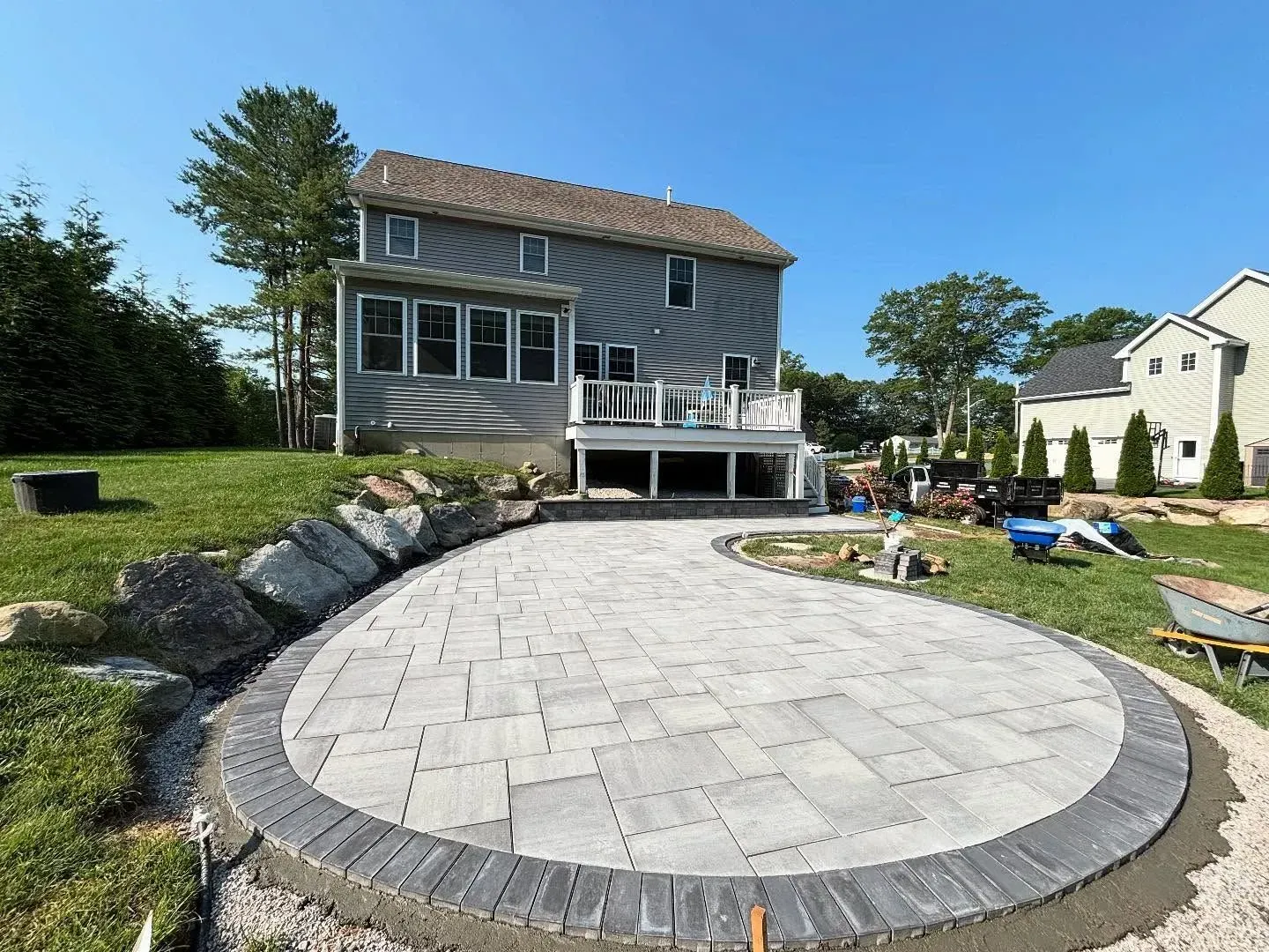 Newly constructed patio with gray pavers and a dark border, adjacent to a house with a deck.