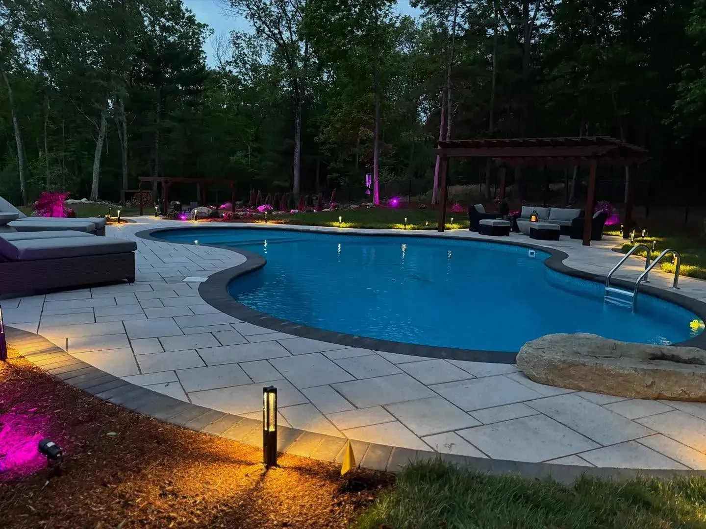 Pool area at dusk with illuminated pathway, pool lights, and pergola.