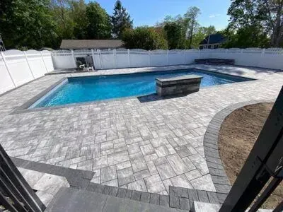 A rectangular swimming pool surrounded by gray pavers, white fence, and greenery under a blue sky.