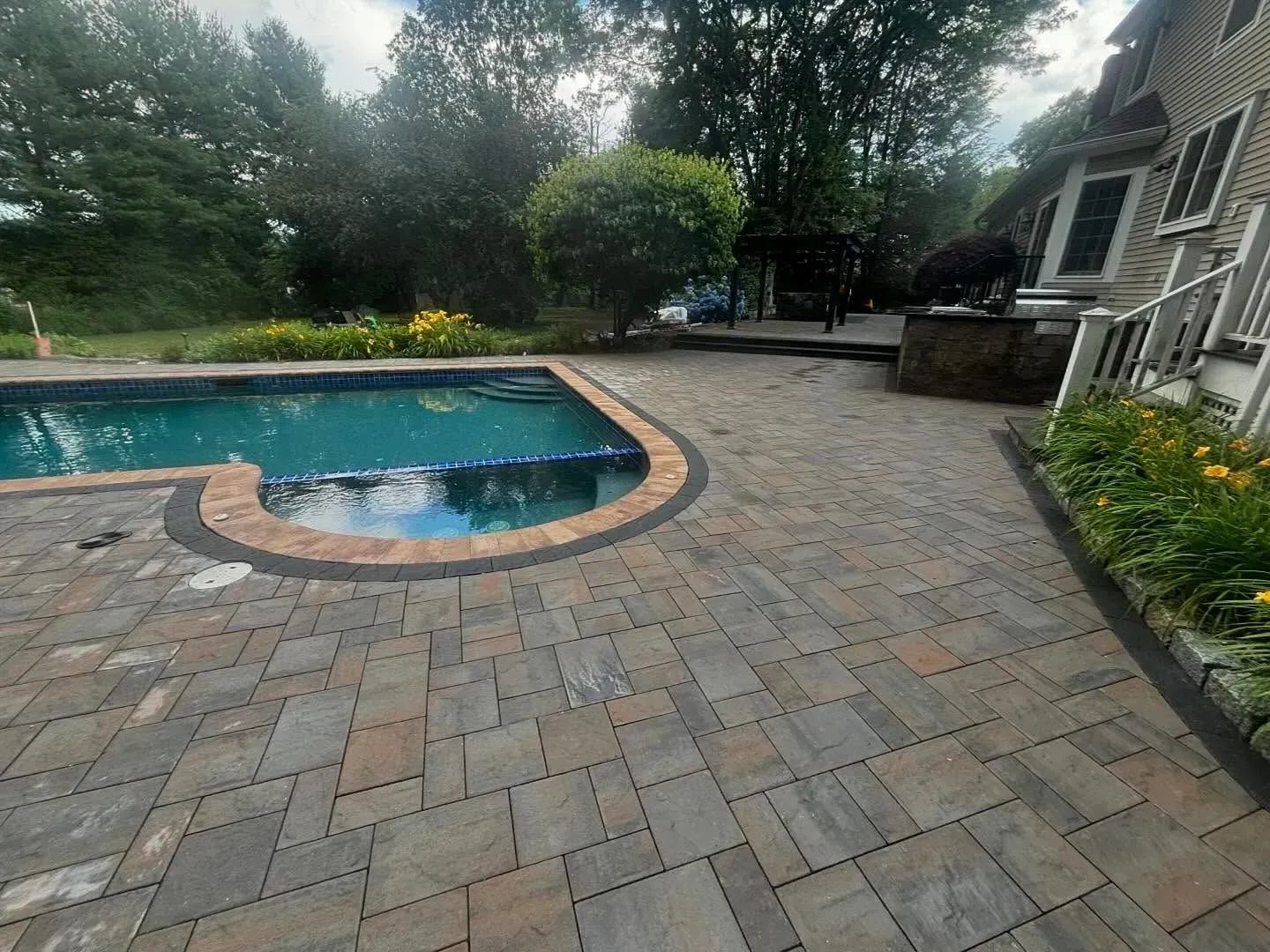Backyard with paved patio, pool, and house. Green trees and yellow flowers visible.