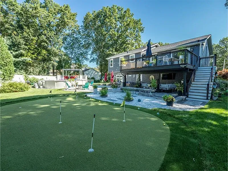 Backyard with putting green, deck, and house. Green lawn, blue sky, and trees.