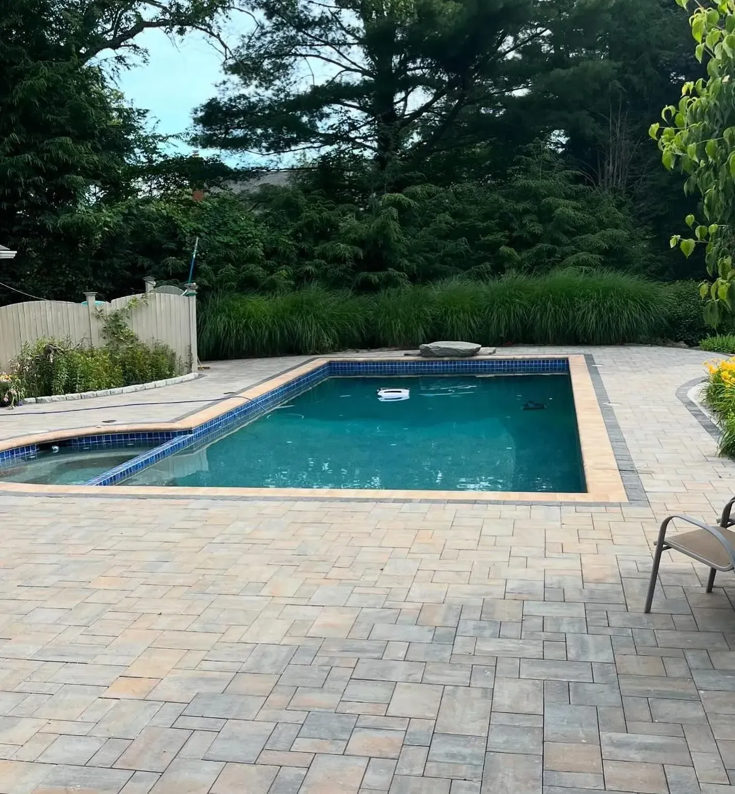 Swimming pool surrounded by stone patio, lush greenery, and a white fence.