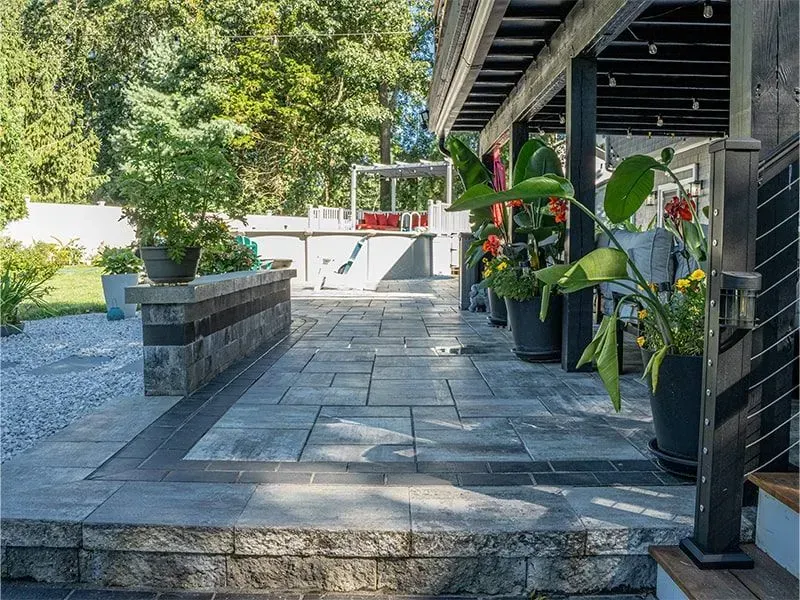 Stone patio with potted plants, low wall, and covered porch. Outdoors with greenery in the background.