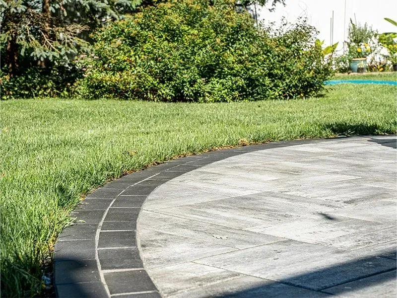Curved gray patio bordered by dark pavers, adjacent to green grass and a bush.