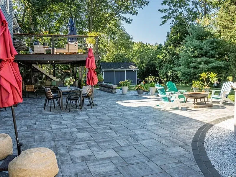 Large paved patio with seating areas, red umbrellas, and a raised wooden deck in a backyard.