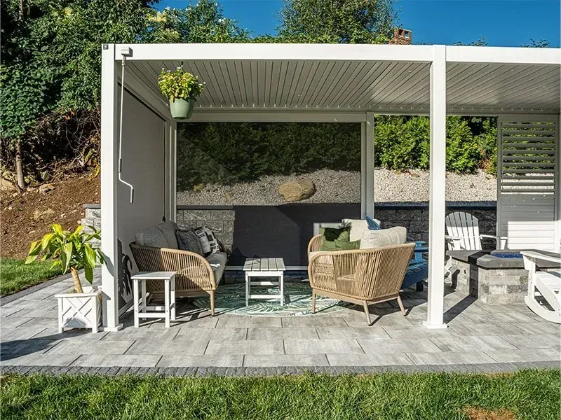 Patio with pergola, wicker furniture, and potted plants on a paved surface, surrounded by greenery.