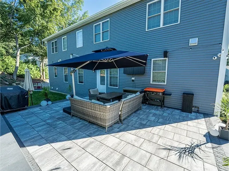Backyard patio with wicker furniture, large umbrella, and grill. Blue house in background.