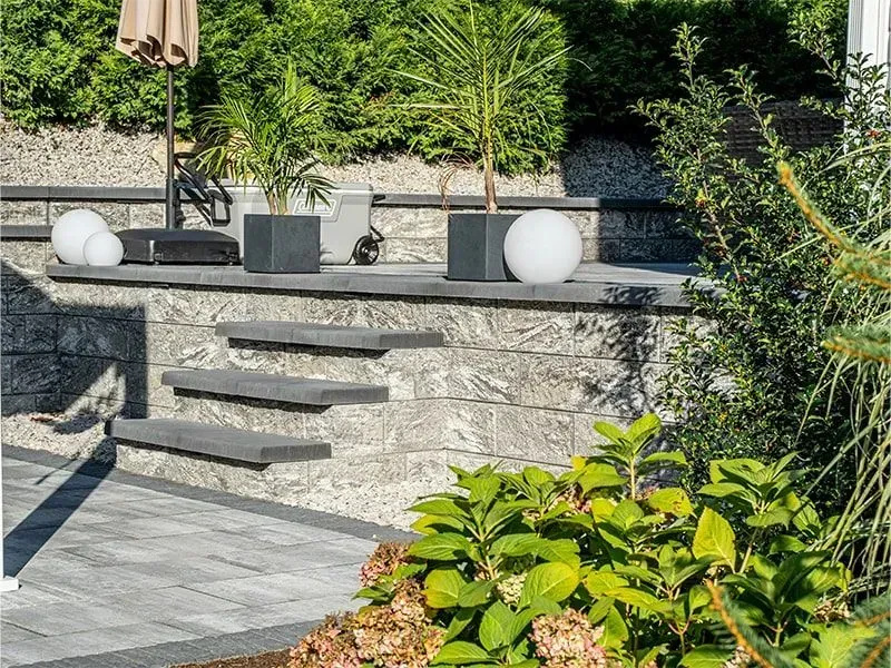 Stone steps lead to a patio with planters, a parasol, and spherical lights, surrounded by greenery.