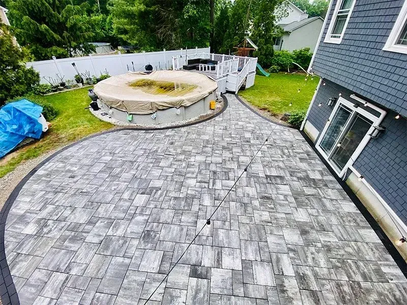 A gray brick patio curves around a pool with a brown cover, connecting to a house and deck.