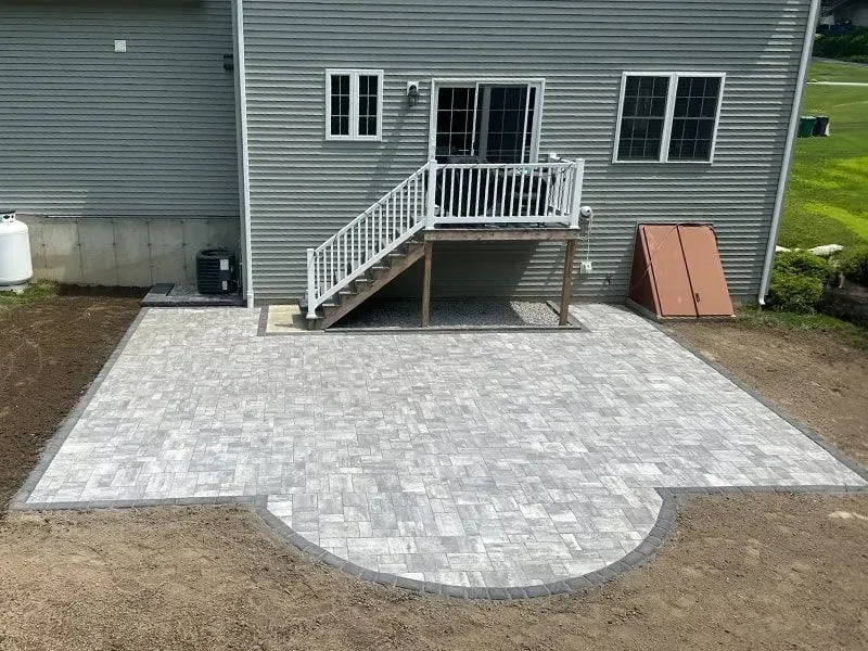 Newly constructed gray paver patio with a curved edge, connected to a house with steps and a door.