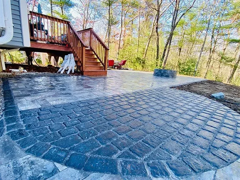 Paver patio with curved design, stairs to a deck, and fire pit in a wooded setting.