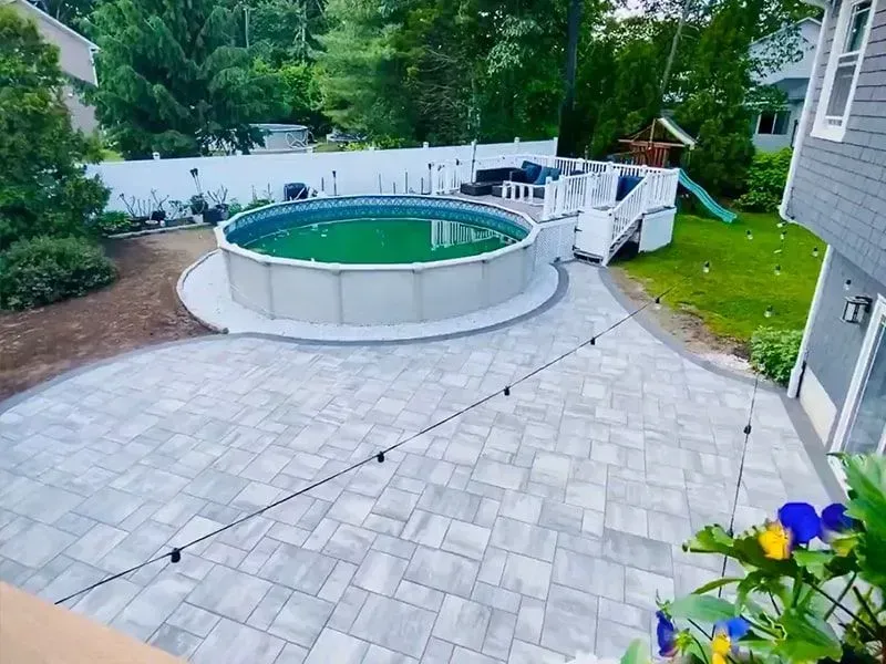 Backyard with an above-ground pool, patio, and white fence. Green grass and trees are visible.