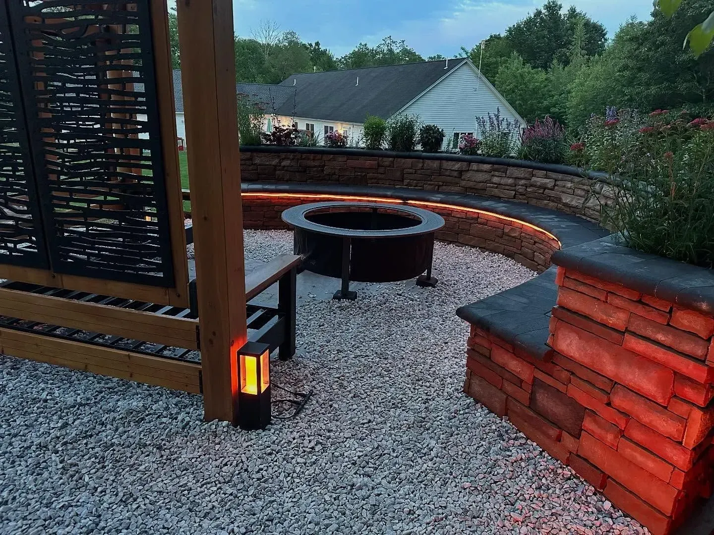 Fire pit in a backyard setting with gravel ground, stone seating, and an illuminated garden feature.
