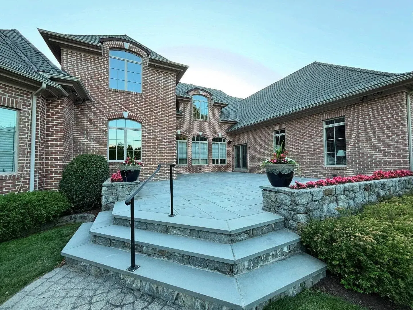 Brick home with stone patio, steps, and black handrail. Planters with flowers.