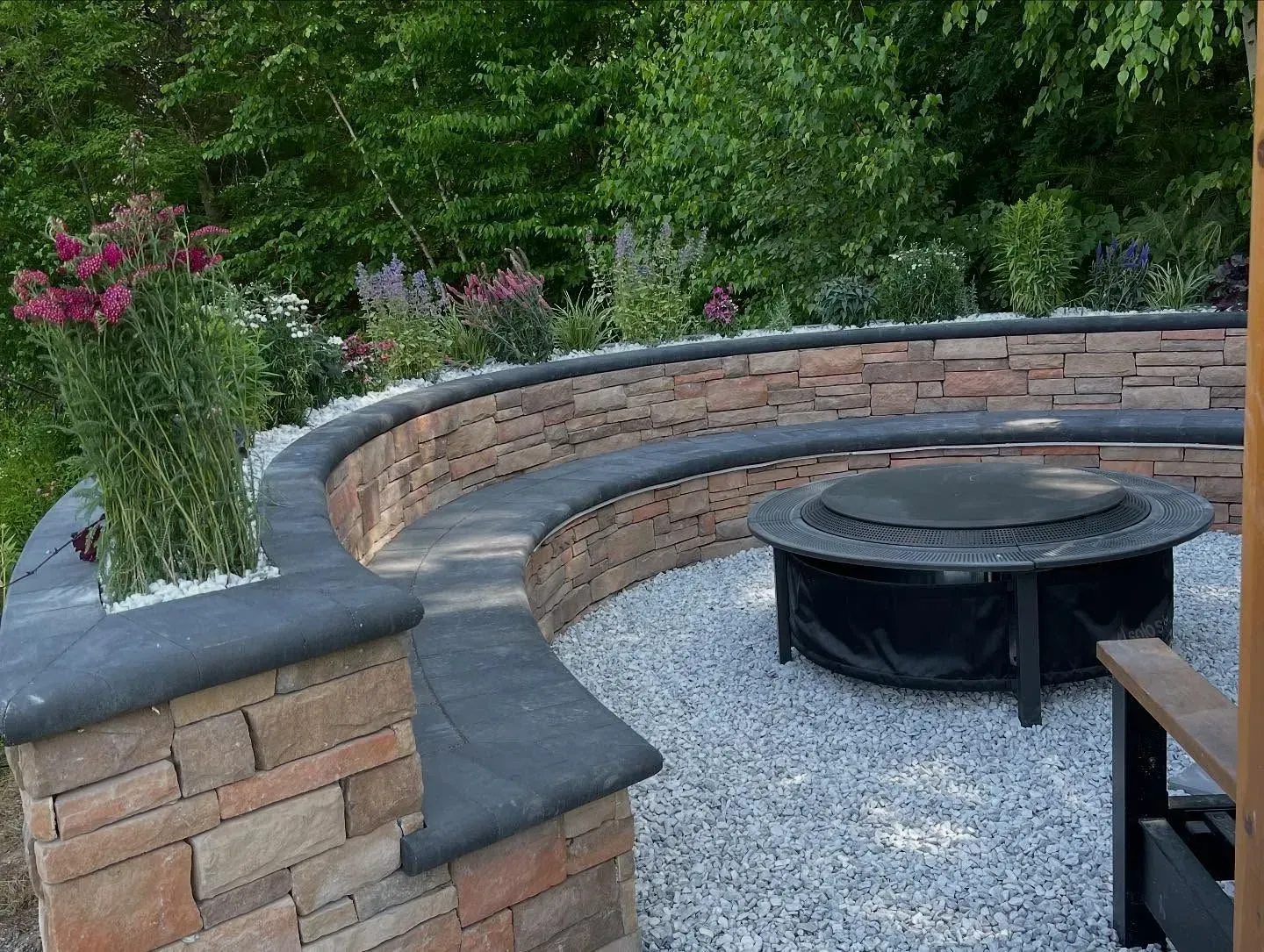 Curved stone wall with built-in seating surrounds a fire pit on a gravel patio with flowering plants.