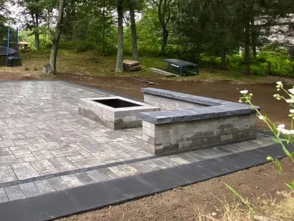 Stone patio with fire pit and seating; surrounded by lawn and trees.