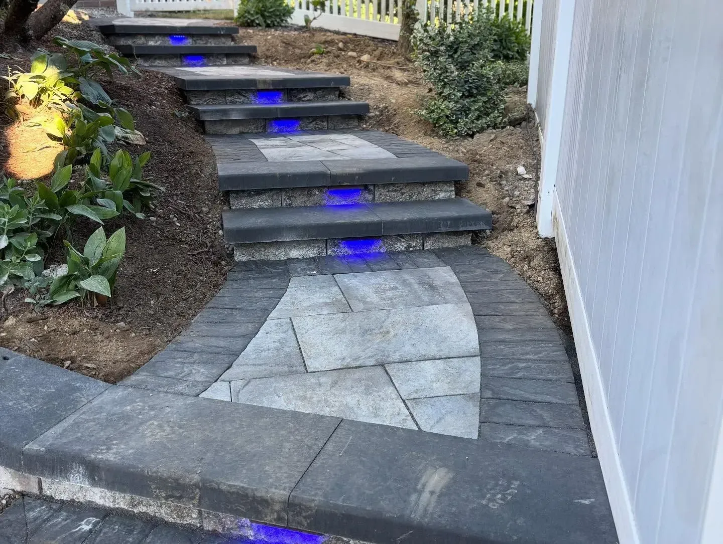 Stone steps with built-in blue lights leading uphill, next to a white fence and garden.