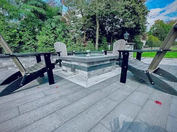 Two Adirondack chairs face a stone fire pit on a paved patio, trees in background.