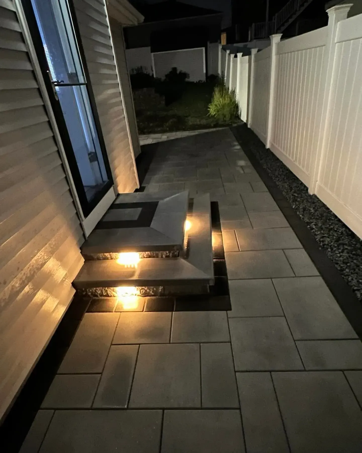 Lit stone pathway and steps leading to a door next to a white fence at night.